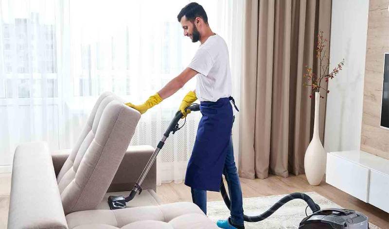 Man in white t-shirt and blue jeans vacuuming sofa inside a living room