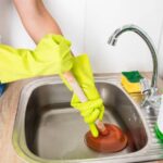 Woman in white top, blue jeans and yellow gloves cleaning drain inside a kitchen