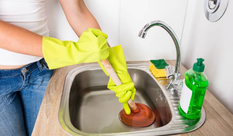 Woman in white top, blue jeans and yellow gloves cleaning drain inside a kitchen