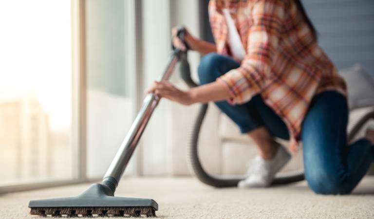 Woman in check shirt and blue jeans vacuuming carpet with vacuum cleaner.