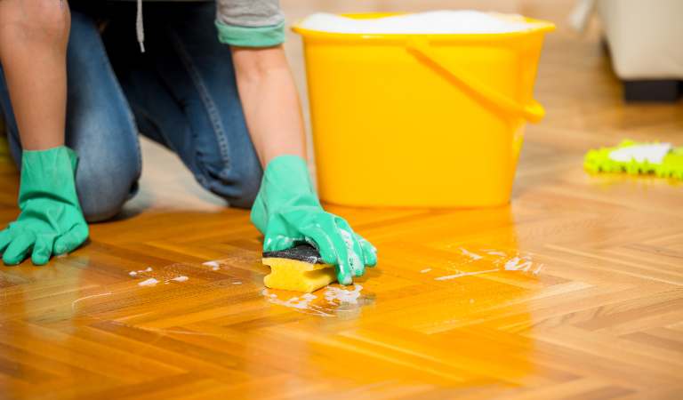 Man in green gloves cleaning floating floor with a yellow brush and yellow basket on the floor.