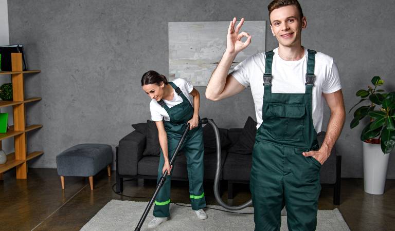smiling young cleaning company workers using vacuum cleaner and showing ok sign