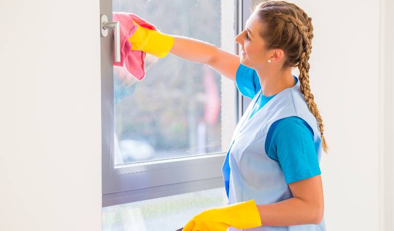 Woman in blue uniform and yellow gloves cleaning window with a pink cloth.