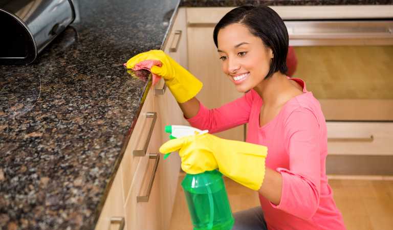 Smiling woman in red top and yellow glove cleaning kitchen cabinet using a green bottle.