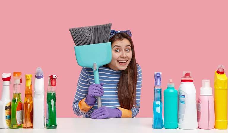 Woman holding a blue cleaning brush with bottles on the table.