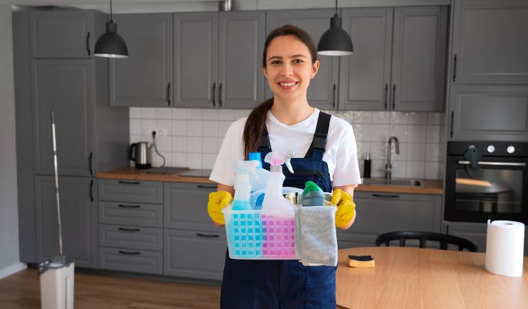 Woman in blue-white uniform holding a basket filled with bottles, tools, cloths.