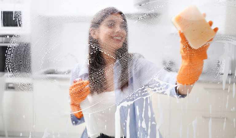 Woman in orange gloves cleaning mirror with hot water and scrubber.