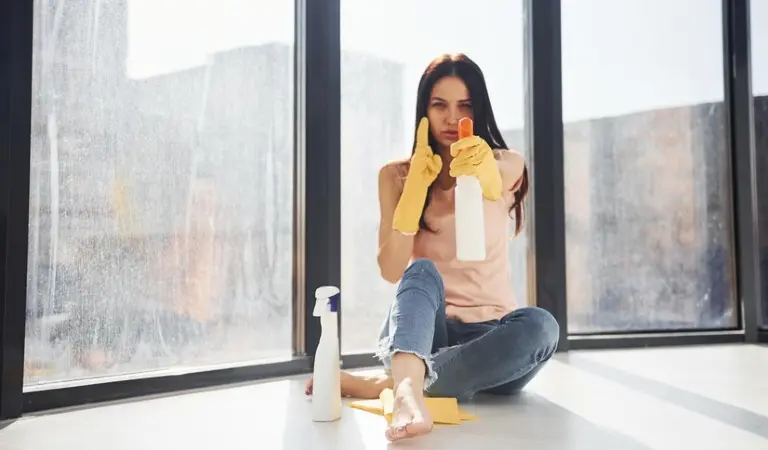 Woman in yellow gloves sitting on floor with a white spray bottle in her hand behind a window