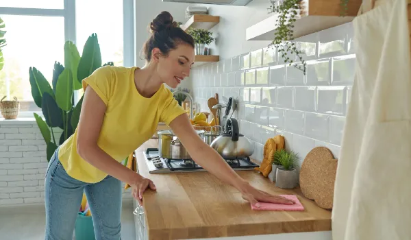 A woman in yellow is cleaning a kitchen counter with a pink towel