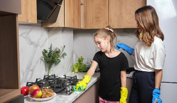 Experienced mother and teen daughter with cleanser in colorful gloves clean marble countertop near stove in light kitchen at home side view