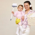 Happy mother housewife is holding cute baby girl with broom in hand and basket in her hand